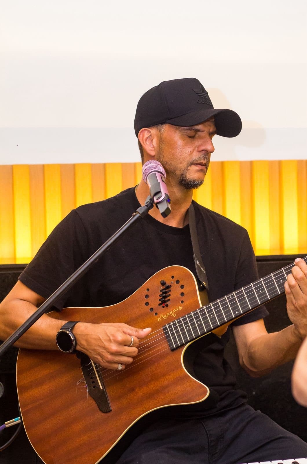 Homem de boné preto toca violão marrom diante de um microfone com fundo amarelo vibrante.