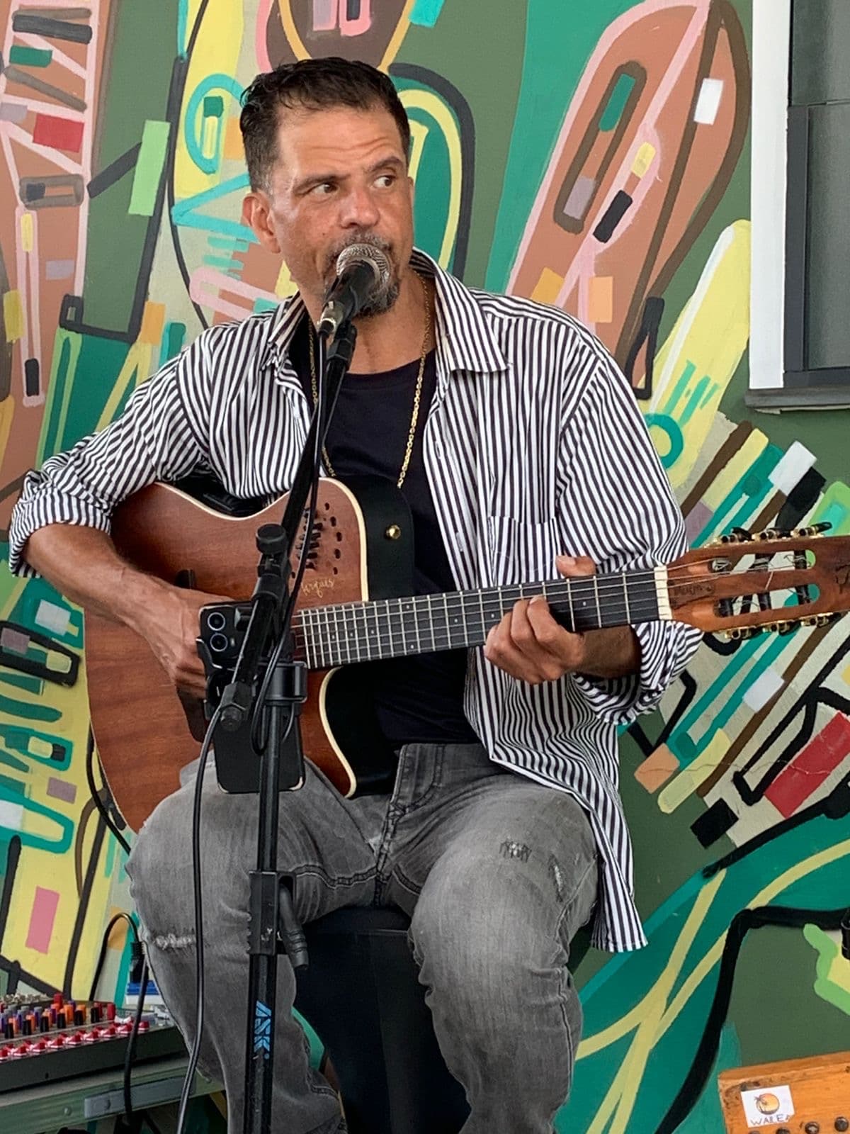 Músico de camisa listrada toca violão e canta em microfone diante de mural colorido.
