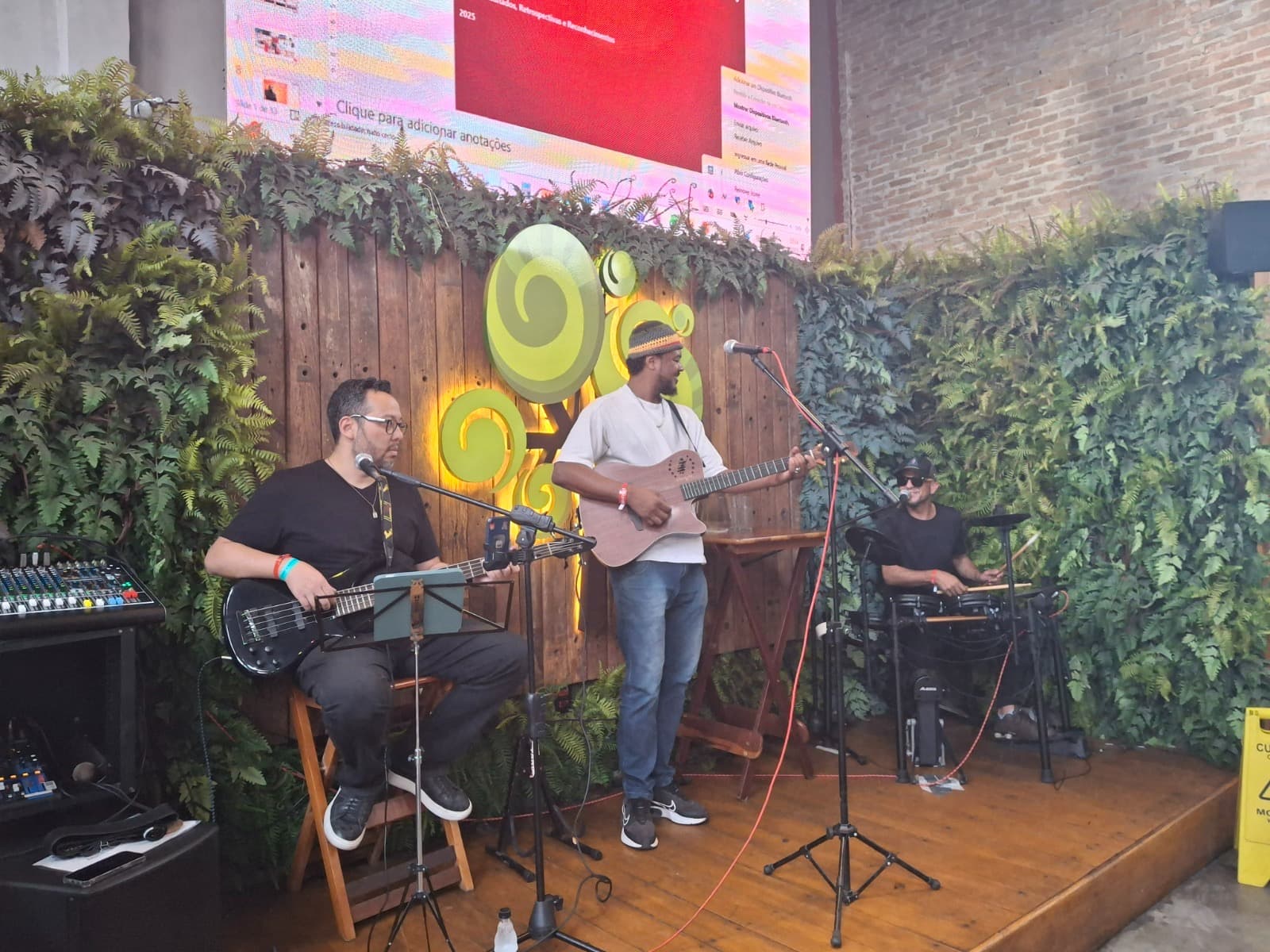 Três músicos tocam baixo, violão e bateria em um palco decorado com plantas verdes.