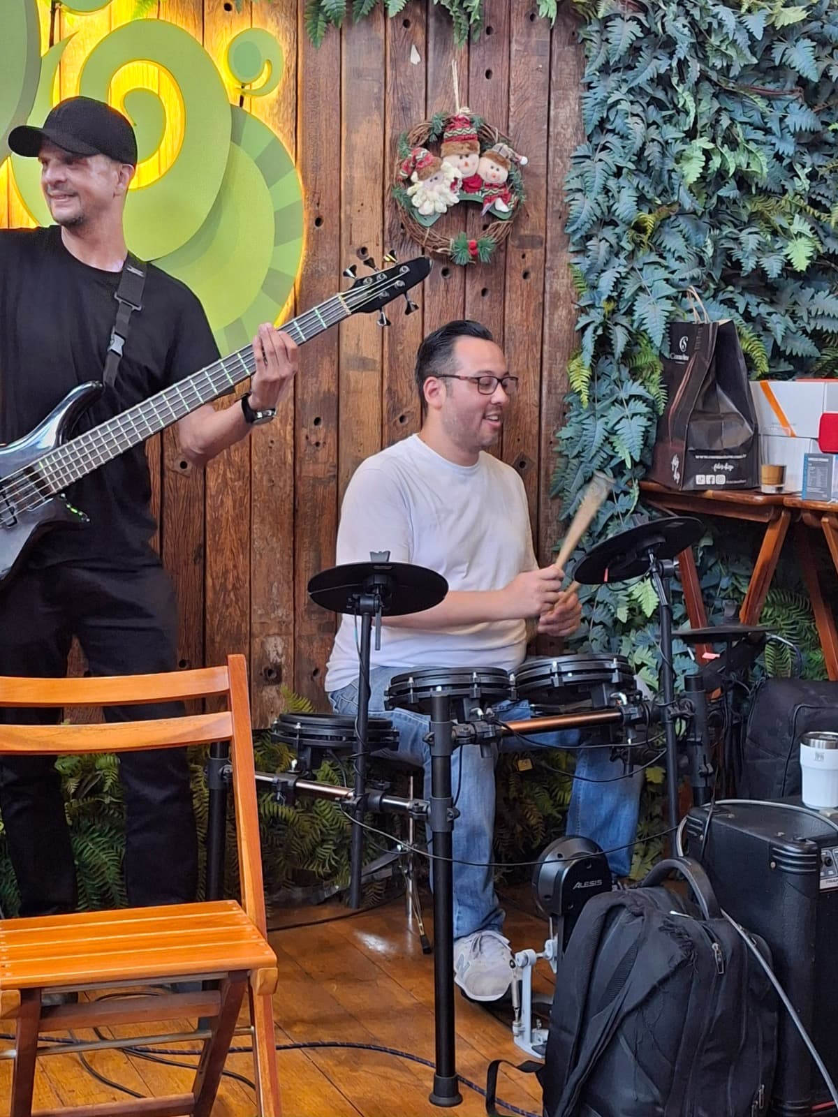 Homem de óculos toca bateria eletrônica ao lado de baixista em palco com fundo amadeirado.