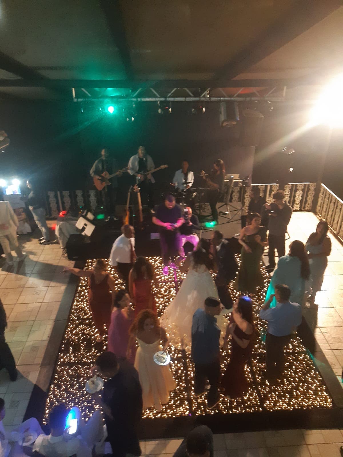 Noiva e convidados dançam em pista iluminada por luzes cintilantes durante festa com banda musical.