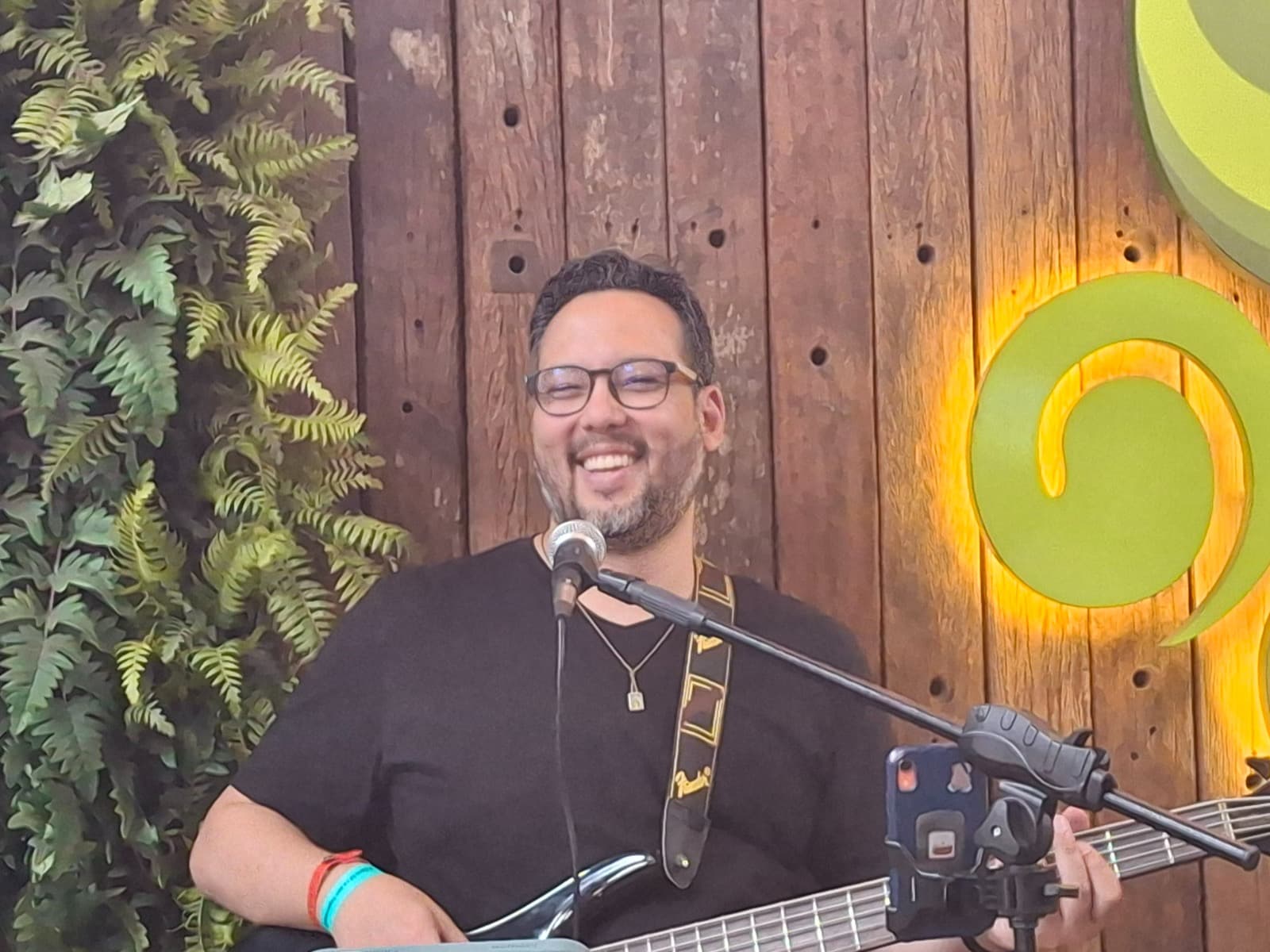Músico sorridente de óculos toca baixo e canta em microfone contra fundo de madeira rústica.