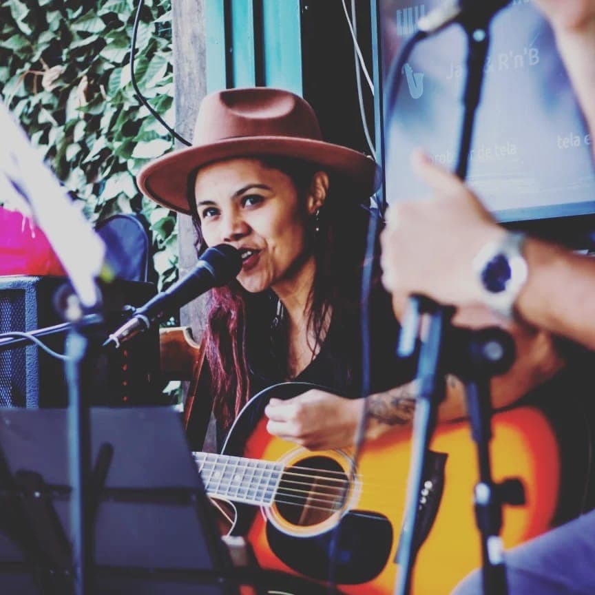 Mulher de chapéu marrom toca violão e canta em um microfone durante uma performance musical.