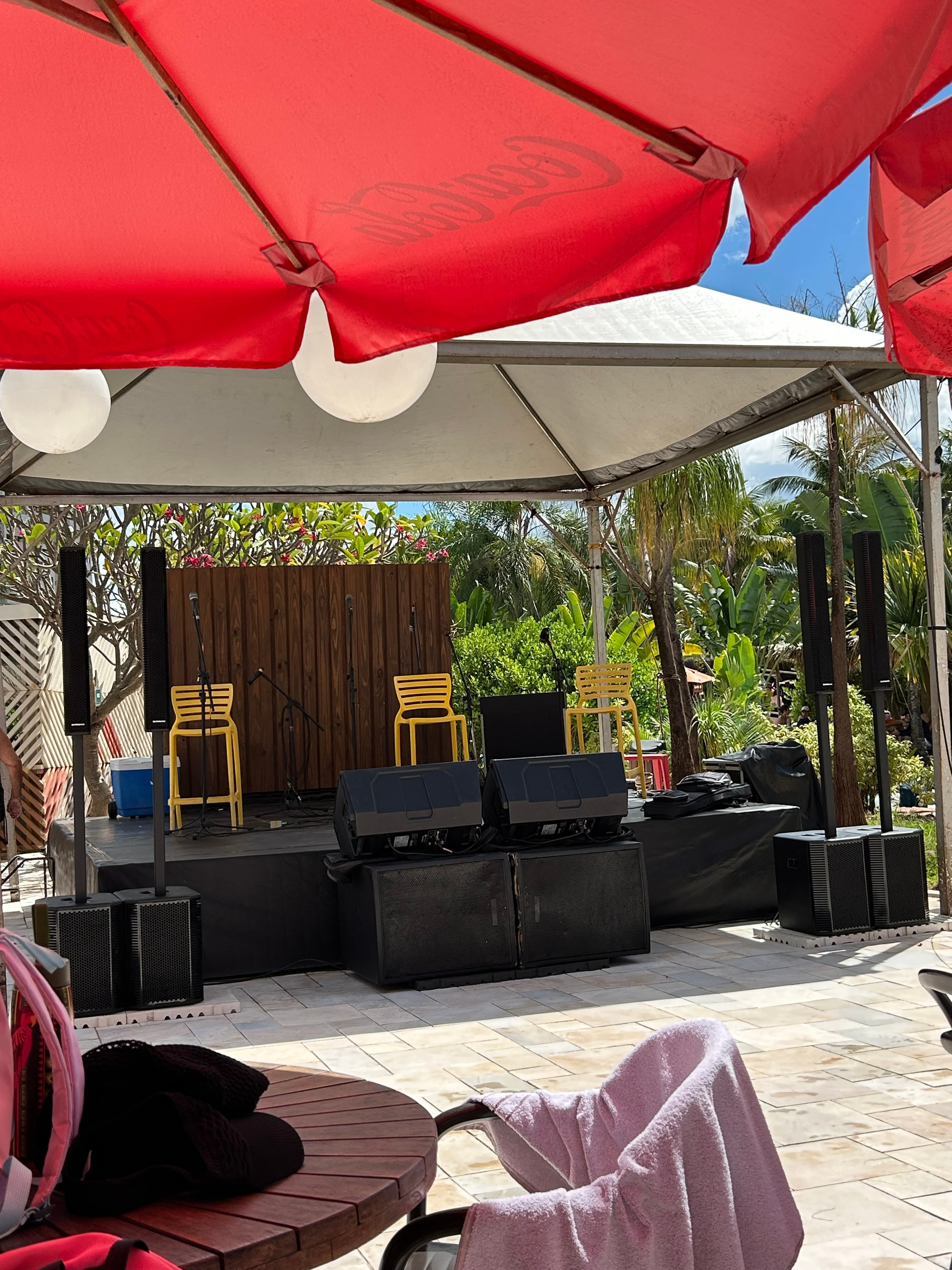 Palco externo com cadeiras amarelas, caixas de som e microfones sob tenda branca e guarda-sol.