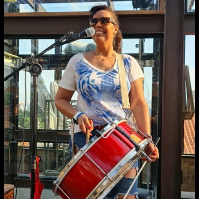 Mulher sorridente de óculos escuros cantando ao microfone e tocando um grande tambor vermelho.