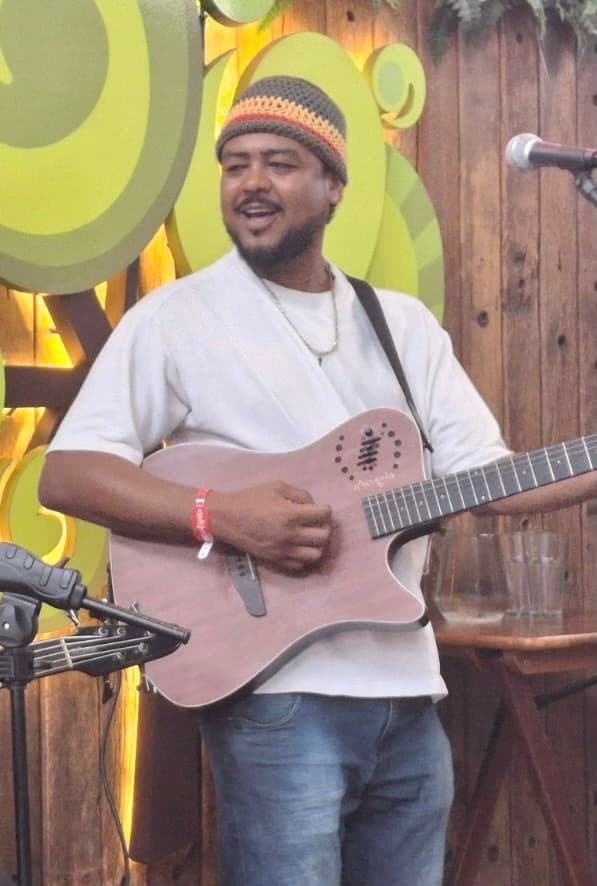 Homem sorridente de touca colorida tocando violão e cantando em um palco rústico de madeira.