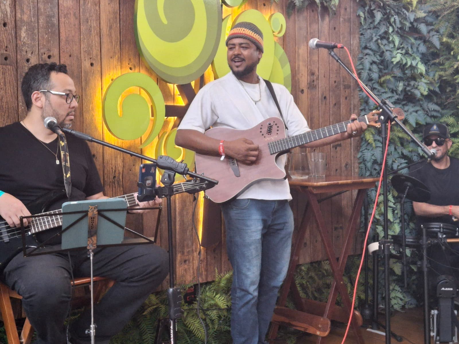 Músicos tocando violão, baixo e bateria em um palco rústico com decorações verdes circulares.
