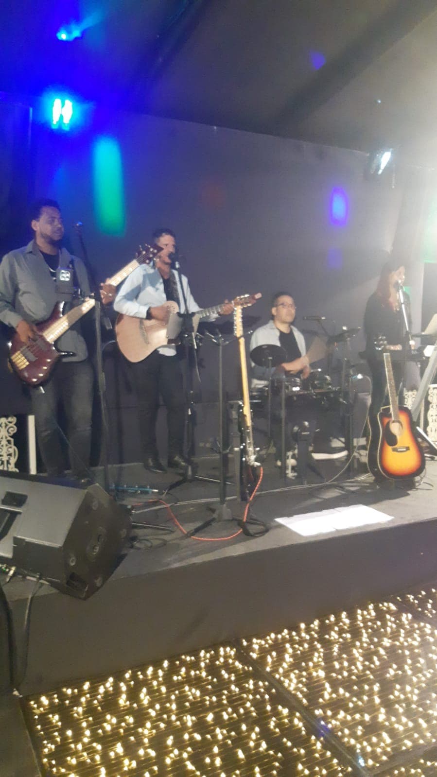 Quatro músicos tocando instrumentos em um palco com luzes coloridas e piso de luzes brilhantes.