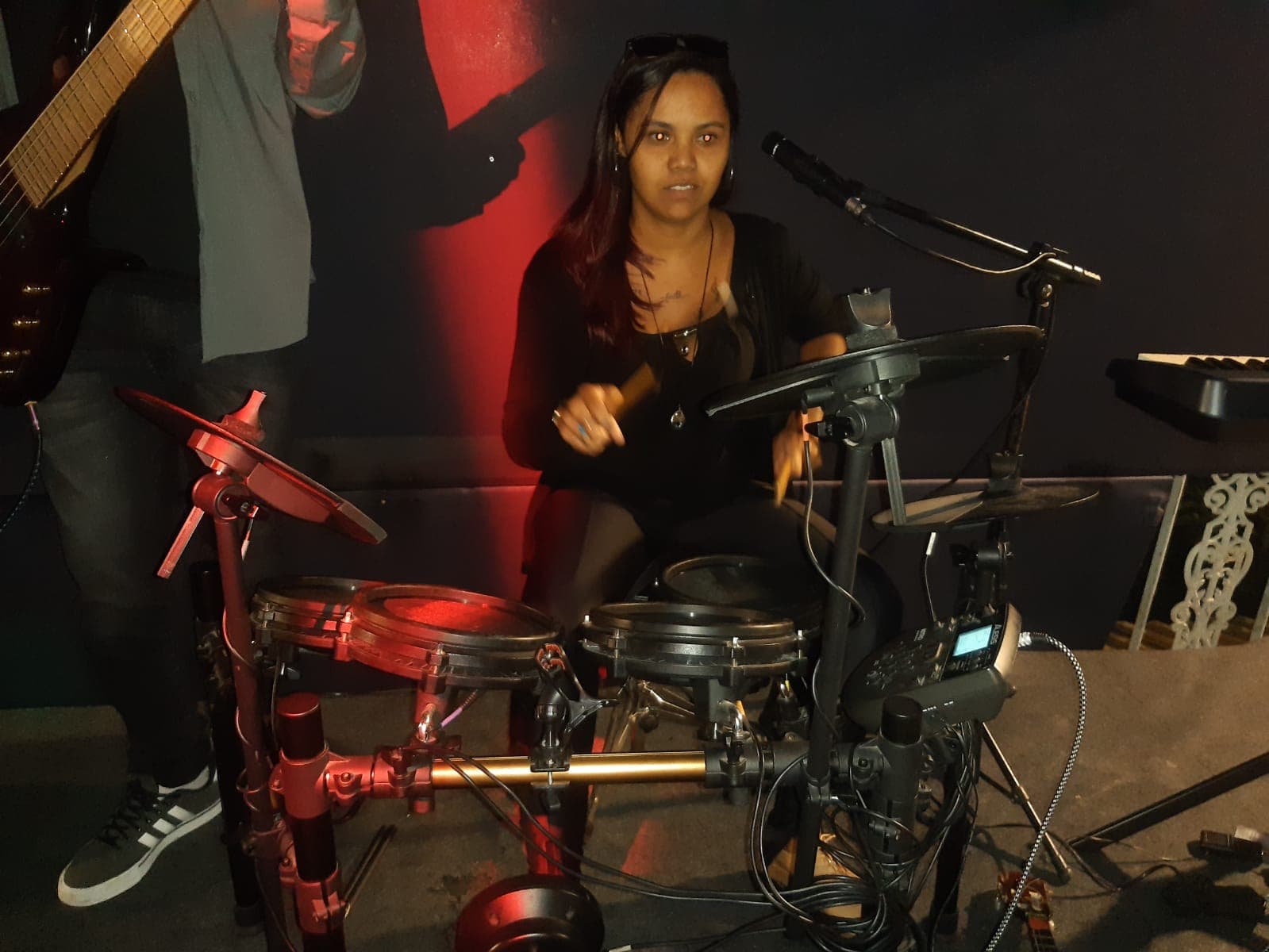 Mulher tocando bateria eletrônica em um palco escuro com iluminação vermelha e baixista ao lado.